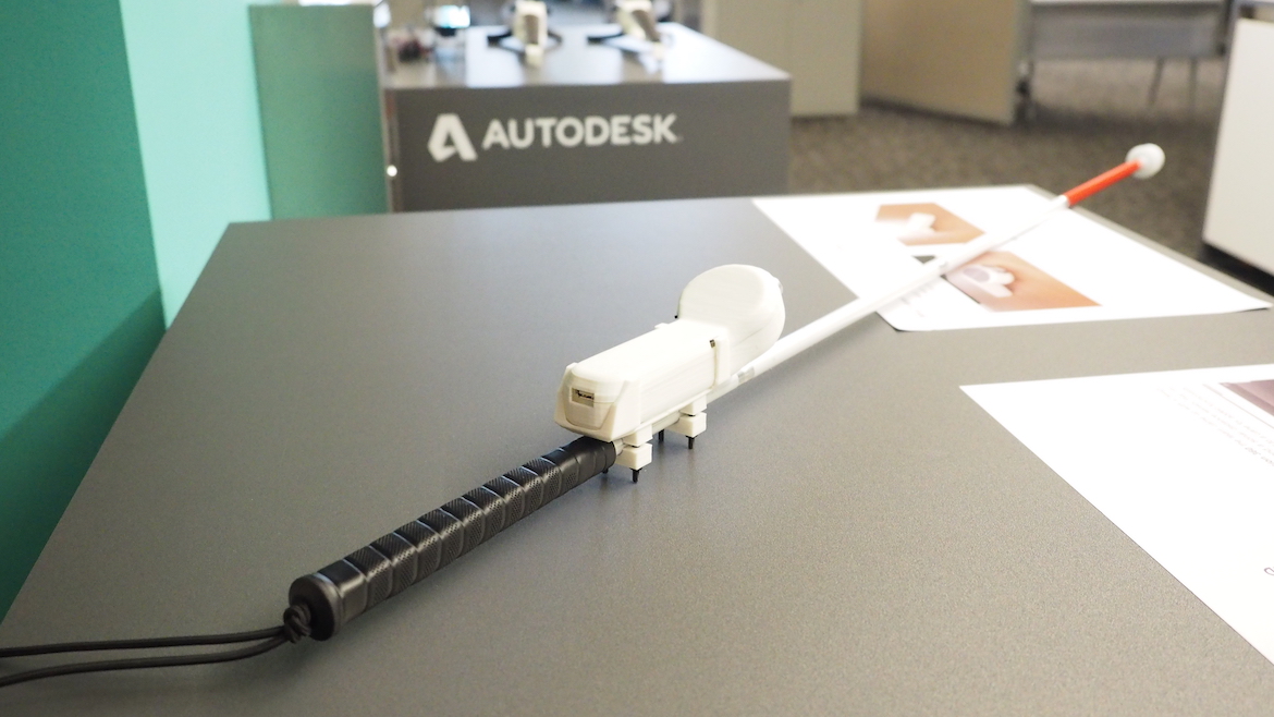 A white cane with a sensor attached to it is displayed on a table. 