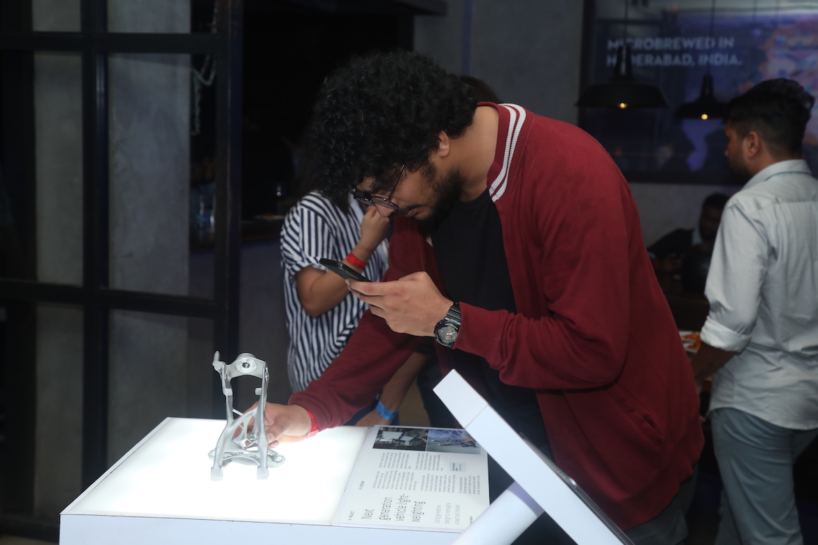 A man takes a photo of a metal 3D-printed seatbelt bracket.