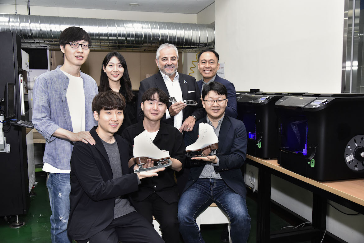 A group of students seated next to a row of 3D printers hold up their new design for a figure skate. The