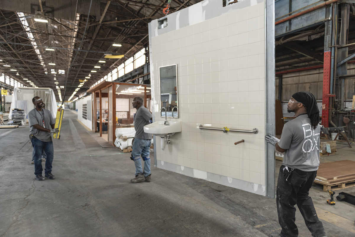 Men transporting a modular wall for a bathroom on a factory floor.