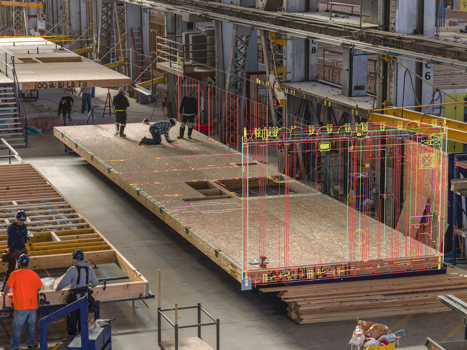 Workers in construction hardhats building modular housing units on a factory floor.