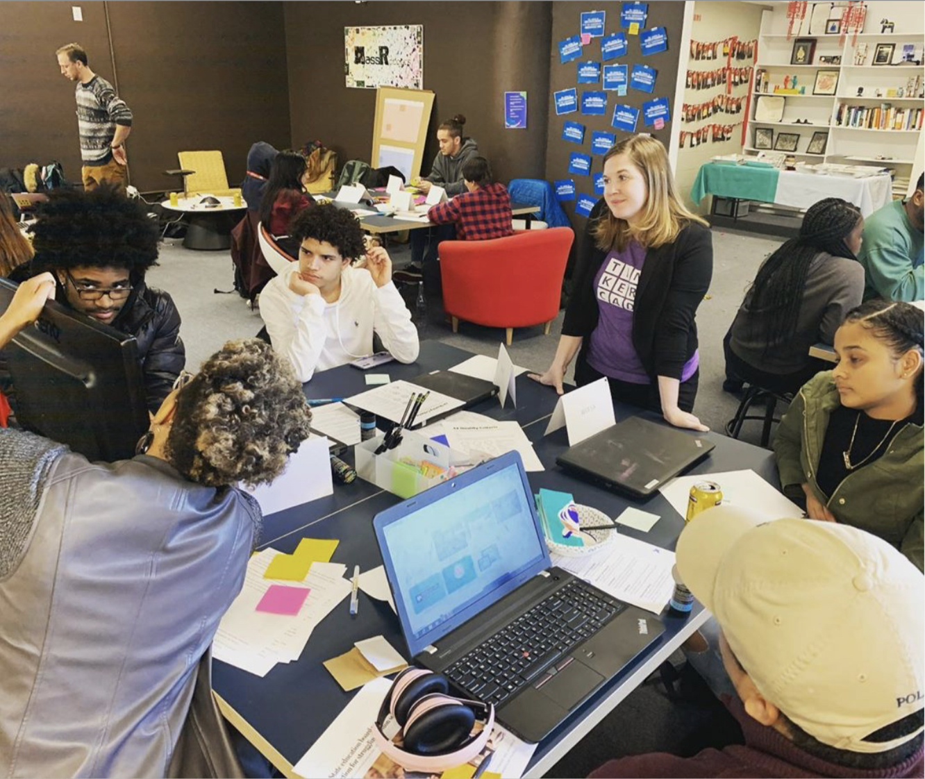 students sitting a table watching other students present as a teacher stands and watches