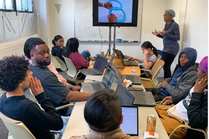 students sitting around a table with laptop computers interacting with a teacher who is standing